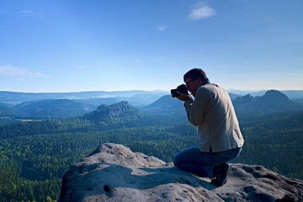 Homem de cabelo escuro está tirando foto por câmera grande espelho no pescoço no pico da montanha ao nascer do sol . — Fotografia de Stock