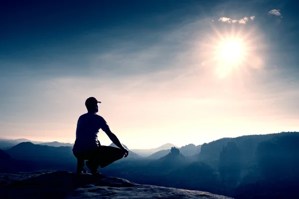 Runner in red cap and  in dark sportswear. Man sit  in squatting position on a rock in heather bushes, enjoy autumn scenery — Stock Photo, Image