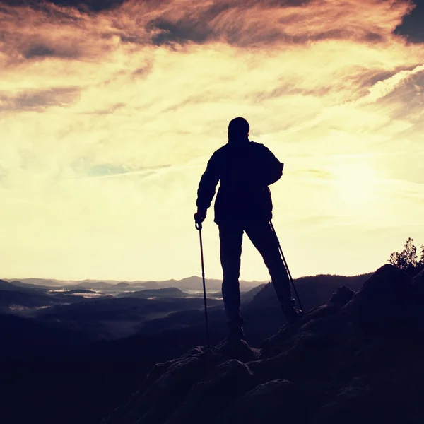 Silhouette of alone hiker with poles in hand. Tourist with sporty backpack stand on rocky view point above misty valley. Sunny spring daybreak in rocky mountains. — Stock Photo, Image