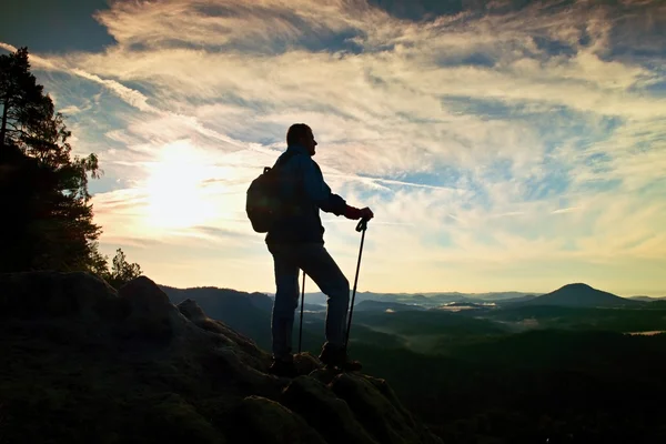 Silueta de excursionista solo con bastones en la mano. Turista con mochila deportiva de pie en el punto de vista rocoso por encima del valle brumoso. Amanecer soleado de primavera en montañas rocosas . — Foto de Stock