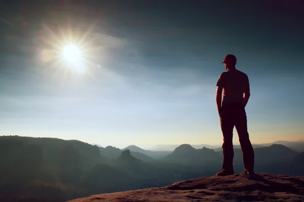 Solo uomo in berretto rosso sulla vetta di picco tagliente nel parco imperi rocciosi e guardando sopra la nebbiosa e nebbiosa valle del mattino al sole. Momento bellissimo — Foto Stock