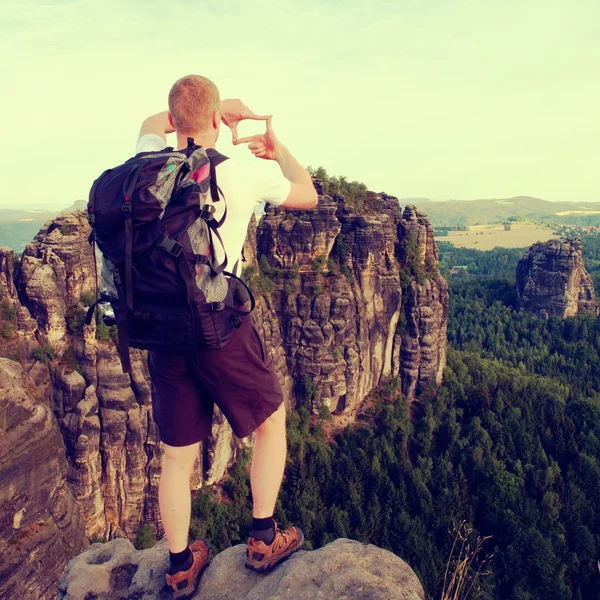 Turista con mochila hacen marco con los dedos en ambas manos. Caminante con gran soporte de mochila en el punto de vista rocoso sobre el bosque . —  Fotos de Stock