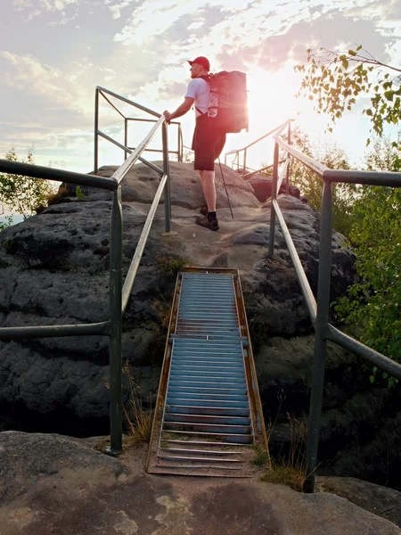 Tall backpacker hålla ledstången på rock. Soliga gryning i klippor. Vandrare med stor ryggsäck, keps, mörka byxor och vit skjorta. — Stockfoto