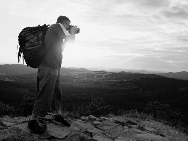 Fotógrafo con una gran cámara de espejo en el cuello y la mochila permanecen en el pico de roca. Paisaje montañoso, color verde fresco en valle . —  Fotos de Stock