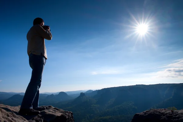 Professional photographer takes photos with mirror camera on cliff of rock. Dreamy misty landscape, hot Sun above — Stock Photo, Image