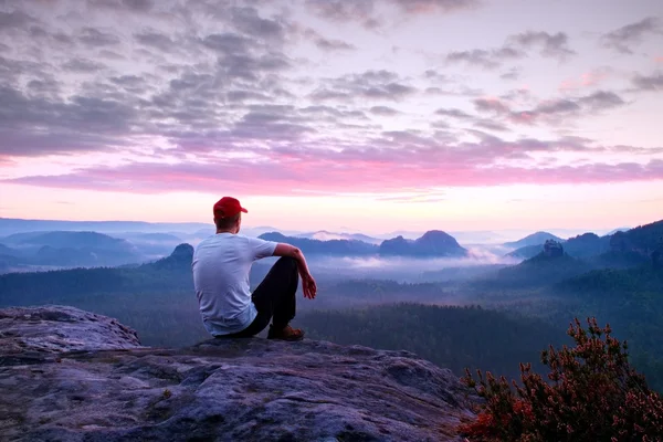 Взрослый спортсмен в белой рубашке, темных брюках и красной кепке. Рыжий короткие волосы человек сидеть на острых скалах над долиной в парке Скалистых гор — стоковое фото