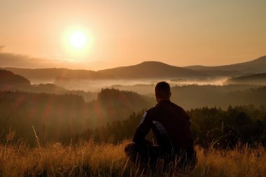 Hiker in squatting position in high grass meadow  enjoy the colorful sunrise scenery
