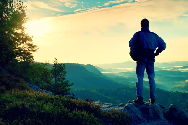 Silhuett av turist med ryggsäck. Soliga våren gryning i Klippiga bergen. Vandrare med sportig ryggsäck stå på rocky synvinkel ovan misty valley. — Stockfoto