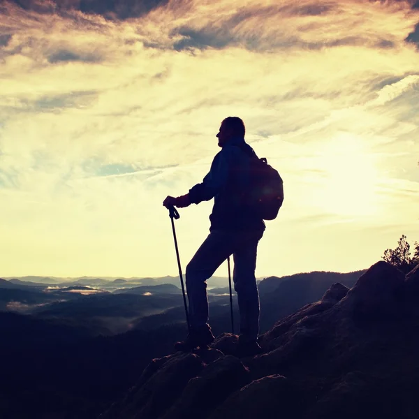 Silhouette of alone hiker with poles in hand. Tourist with sporty backpack stand on rocky view point above misty valley. Sunny spring daybreak in rocky mountains. — Stock Photo, Image