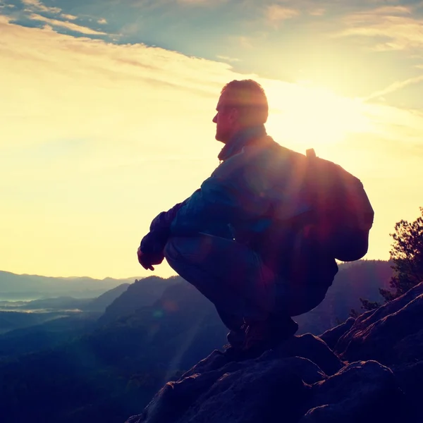 Senderista con mochila deportiva sentarse en el borde del acantilado rocoso y ver el valle brumoso a continuación. Amanecer soleado de primavera en montañas rocosas . — Foto de Stock