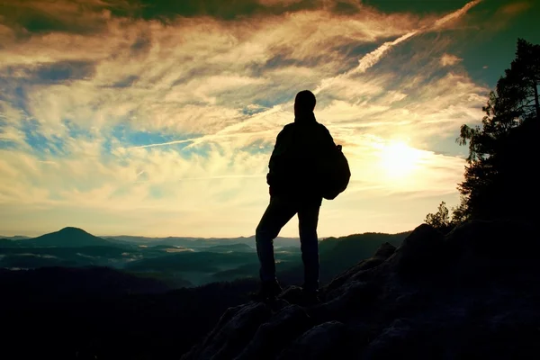 Turista con mochila deportiva de pie en el punto de vista rocoso y mirando en profundo valle brumoso a continuación. Amanecer soleado de primavera en montañas rocosas . — Foto de Stock