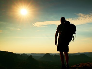 Tall tourist with poles in hand. Sunny evening in rocky mountains. Hiker with big backpack stand on rocky view point above misty valley. Vignetting effect.