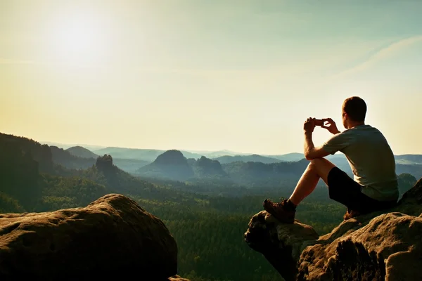 Turista en camiseta gris toma fotos con el teléfono inteligente en el pico de roca. Paisaje de ensueño colinas abajo, naranja rosado amanecer brumoso en un hermoso valle por debajo de las montañas rocosas. — Foto de Stock