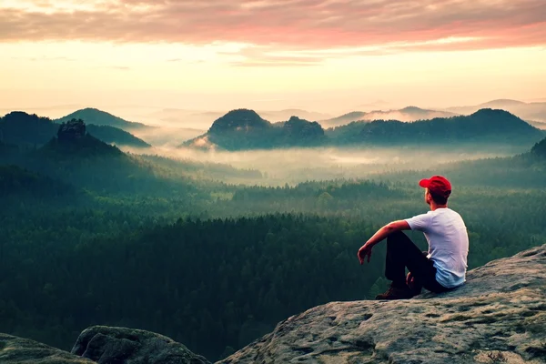 Adult sportsman in white shirt, dark trousars and red cap. Ginger short hair man sit on sharp cliff above valley in rocky mountains park — Stock Photo, Image