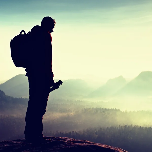 Alone  photographer with heavy backpack and tripod in hand on rocky cliff and watching down to deep misty valley bellow — Stock Photo, Image