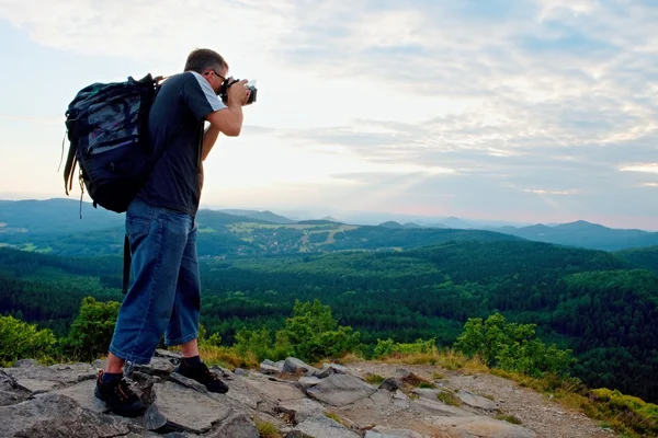 Fotograf mit großer Spiegelkamera am Hals und Rucksack bleiben auf dem Gipfel des Felsens. Hügelige Landschaft, frische grüne Farbe im Tal. — Stockfoto