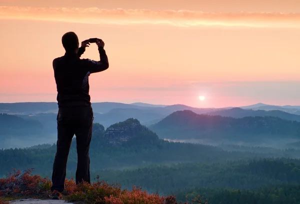 Man takes photos with smart phone on peak of rock empire. Dreamy fogy landscape, spring orange pink misty sunrise above beautiful valley — Stock Photo, Image