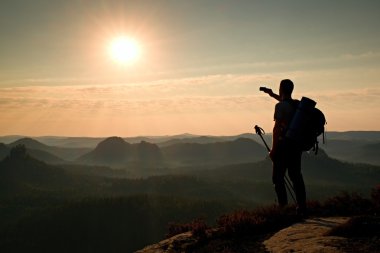 Tall backpacker with poles in hand. Sunny daybreak in mountains. Hiker with big backpack on rocky view point above misty valley.