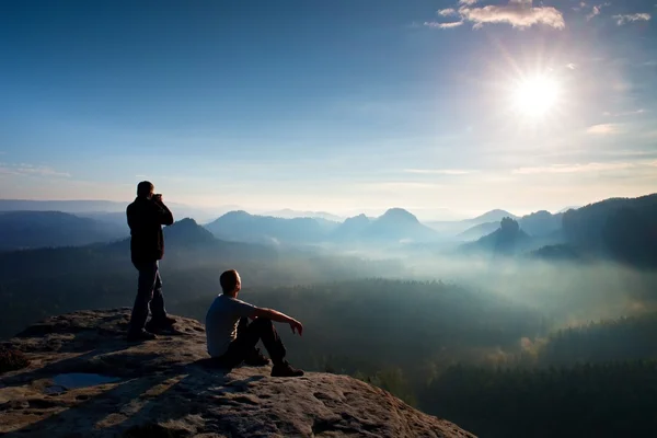 Turysta i entuzjastów fotografii zatrzymać ze statywem na klif i myślenia. Marzycielski fogy krajobraz, niebieski mglisty wschód słońca w pięknej dolinie poniżej — Zdjęcie stockowe
