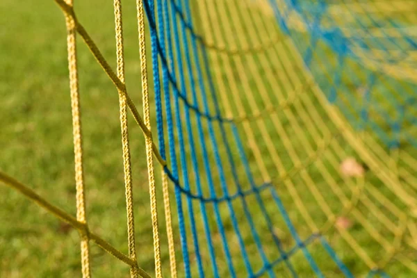 Detail der gelb blau gekreuzten Fußballnetze, Fußball im Tornetz mit schlechtem Gras auf dem Spielplatz im Hintergrund. — Stockfoto