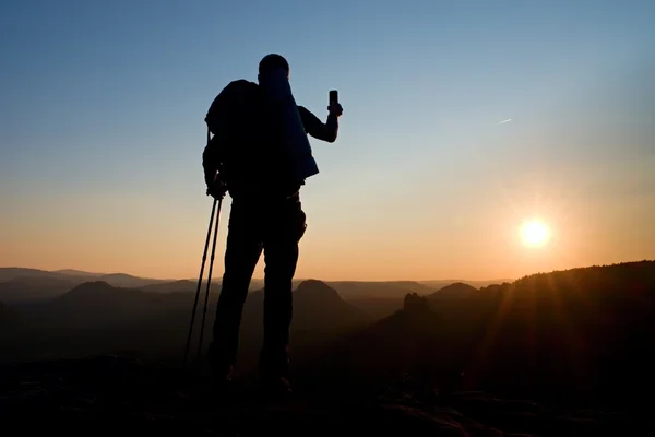 Турист с большим рюкзаком фотографируется со смартфоном на скалистом пике. Мечтательная туманная долина внизу — стоковое фото