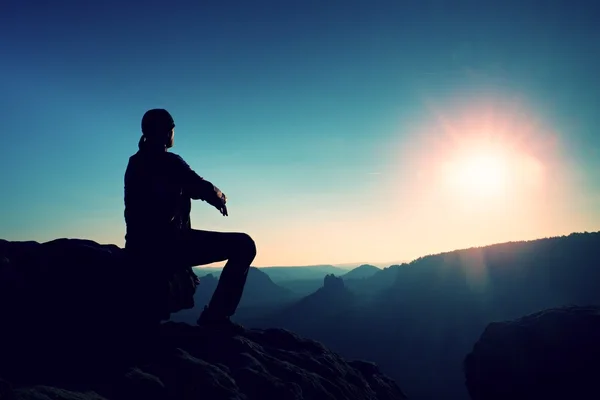 Young man in black sportswear is sitting on cliff and looking to misty valley bellow — Stock Photo, Image