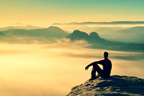 Hiker in black on the rocky peak. Wonderful daybreak in mountains, heavy orange mist in valley. Man sit on the rock. — Stock Photo, Image