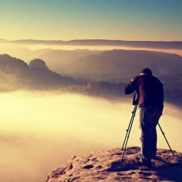 Professionista su Cliff. Fotografo naturalistico scatta foto con macchina fotografica a specchio su roccia. Paesaggio nebbioso da sogno, nebbia rosa arancio primaverile sotto . — Foto Stock