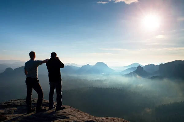 Dos amigos. Caminante pensando y entusiasta de la foto toma fotos estancia en el acantilado. Paisaje de ensueño fogy, azul brumoso amanecer en un hermoso valle debajo — Foto de Stock