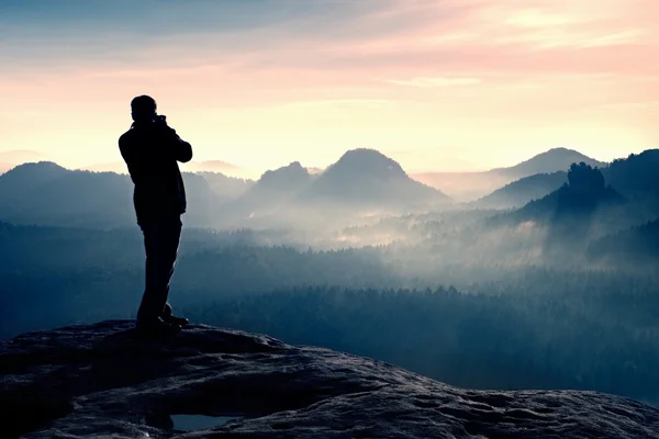 Fotógrafo profissional tira fotos com câmera grande no rock. Possibilidade perigosa no fim do penhasco. Paisagem nebulosa sonhadora, sol quente acima — Fotografia de Stock