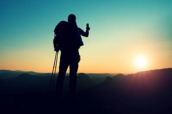 Wanderer mit großem Rucksack fotografiert mit dem Smartphone auf dem felsigen Gipfel. verträumtes Nebeltal unten — Stockfoto