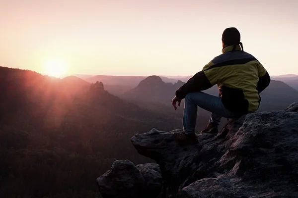 Visão traseira do caminhante masculino em casaco preto amarelo sentado em penhasco rochoso enquanto desfruta do amanhecer acima do vale — Fotografia de Stock