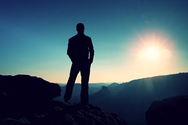 Slim turista en el pico agudo de la montaña rocosa está vigilando el valle de la mañana brumoso y nebuloso al sol — Foto de Stock