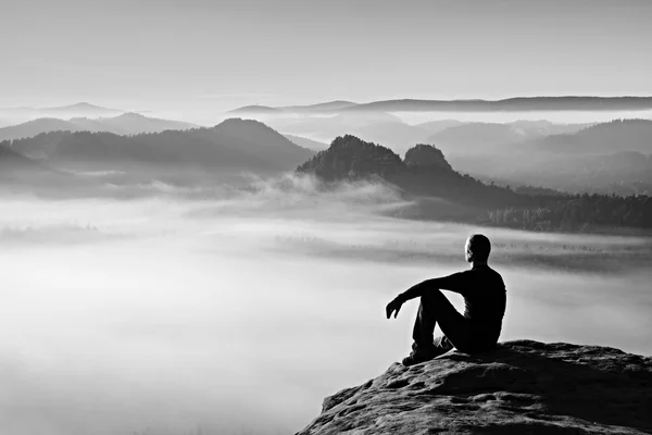 Caminante en negro en el pico rocoso. Maravilloso amanecer en las montañas, niebla naranja pesada en el valle. El hombre se sienta en la roca . —  Fotos de Stock