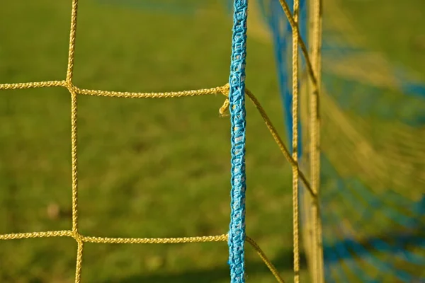Detail der gelb blau gekreuzten Fußballnetze, Fußball im Tornetz mit schlechtem Gras auf dem Spielplatz im Hintergrund. — Stockfoto