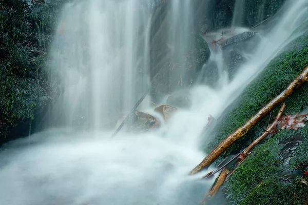 Spray de água abaixo pequena cachoeira no córrego da montanha, a água está caindo sobre pedra musgosa. O spray criar no nível e cascalho de água leitosa. Ramos e troncos partidos na água — Fotografia de Stock