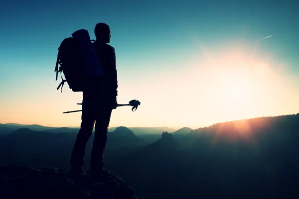 Hoog backpacker met palen in de hand. Wandeling van de zonnige dag in de rocky mountains. Wandelaar met grote rugzak staan op rotsachtige oogpunt boven mistige vallei. — Stockfoto