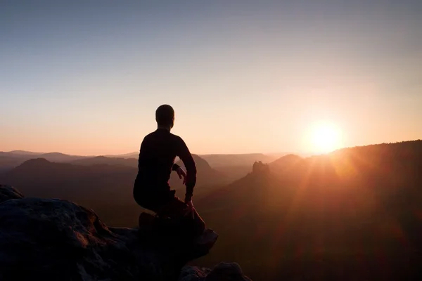 Joven en ropa deportiva negro sentarse en el borde del acantilado y mirar en el amanecer en el horizonte sobre el valle brumoso — Foto de Stock