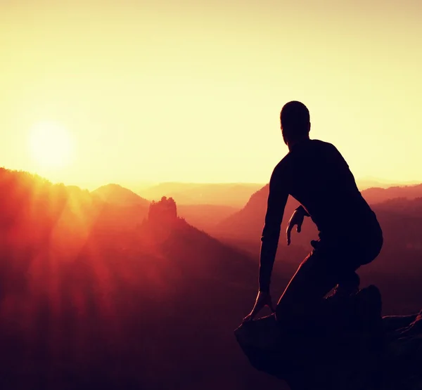 Joven en ropa deportiva negro sentarse en el borde del acantilado y mirar en el amanecer en el horizonte sobre el valle brumoso — Foto de Stock