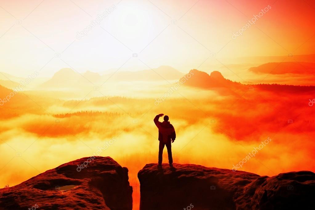 Beautiful moment the miracle of nature. Man stands on the peak of sandstone rock in national park Saxony Switzerland and watching over the misty and foggy morning valley to Sun. 