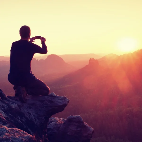 Young man in black sportswear is sitting on sharp cliff and taking photos by smart phone — Stock Photo, Image