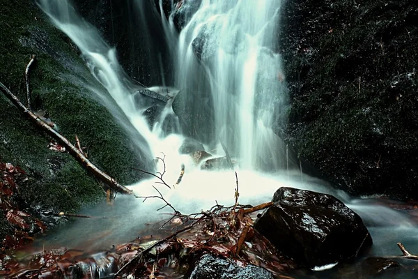 Водный спрей под небольшим водопадом на горном ручье, вода падает над мшистым валуном. Спрей создает на уровне и щебень молочной воды. Сломанные ветви в воде — стоковое фото