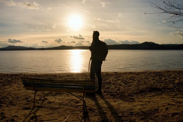 Alone traveler with backpack. Man  on sea beach at wooden bench, cold sunny autumn evening — Stock Photo, Image
