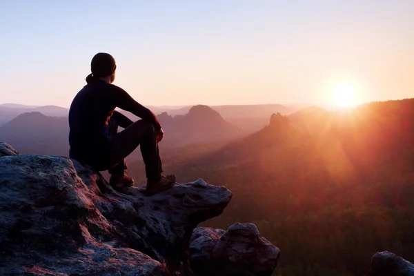 Tired adult hiker in black trousers, jacket and dark cap sit on cliff edge and looking to colorful mist in valley bellow — Stock Photo, Image