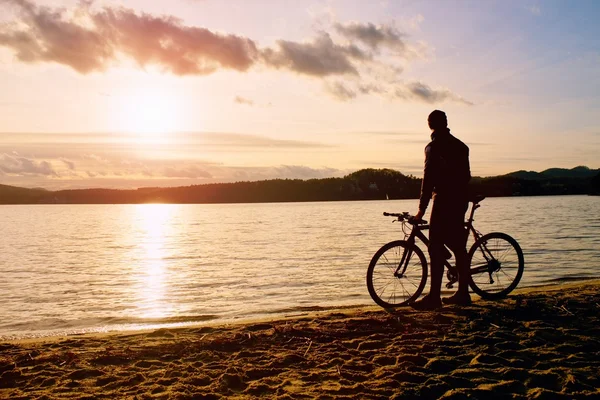 Joven ciclista silueta en el cielo azul y puesta de sol sobre la playa. Motociclista al final de la temporada en el lago . — Foto de Stock