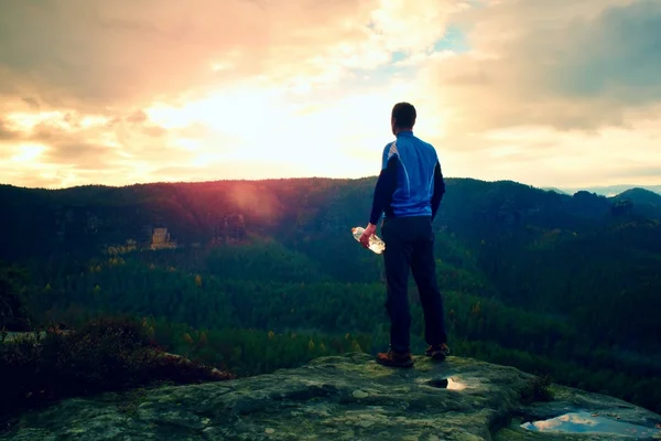 Svettig tall vandrare i cyan trikot med flaska vatten. Vuxen trött turist på toppen av rock watch in dimmiga landskap. — Stockfoto