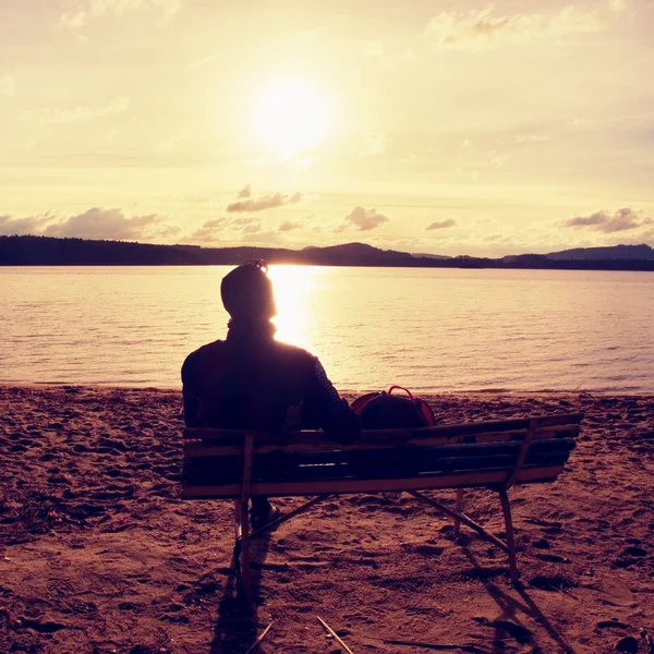 Sam młody człowiek w sylwetka siedzi w słońcu na plaży. Turystyczny odpocząć na ławeczce, drewniane, na jesień jezioro. — Zdjęcie stockowe