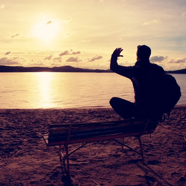 Seul jeune homme en silhouette assis au soleil sur la plage. Touriste se reposer sur banc en bois au lac d'automne . — Photo