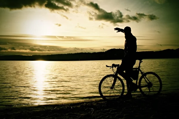 Silhueta de desportista segurando bicicleta no lago bech, colorido céu nublado por sol e reflexão no nível ondulado da água — Fotografia de Stock