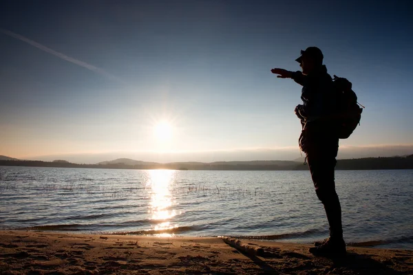 Homme avec la main en l'air. Grand randonneur en vêtements de sport sombres avec des stands de sac à dos sportif sur la plage profiter du coucher de soleil à l'horizon — Photo